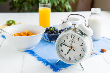 Time for breakfast. Alarm clock with healthy food on the background. Bowl with cereals, nuts and berries, milk, fresh juice on blue napkin and white wooden table. Day diet planning and healthy eating.