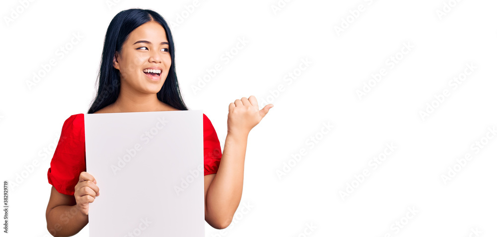 Young beautiful asian girl holding blank empty banner pointing thumb up to the side smiling happy with open mouth