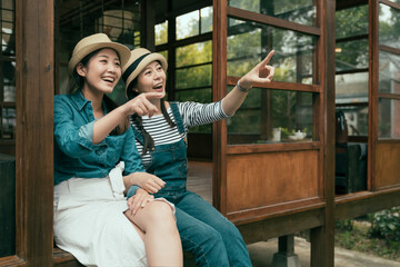 Wall Mural - two cheerful young asian chinese girl travelers pointing finger away while relax sitting on wooden floor. friends in straw hats smiling enjoy garden view from japanese traditional design house.