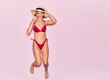 © Krakenimages.com - Young beautiful girl on vacation wearing bikini and summer hat smiling happy. Jumping with smile on face doing victory sign over isolated pink background