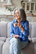 © insta_photos - Happy beautiful relaxed mature older adult grey-haired woman drinking coffee relaxing on sofa at home. Smiling stylish middle aged 60s lady enjoying resting sitting on couch in living room, vertical.