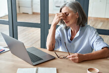 Overworked Tired Older Lady Holding Glasses Feeling Headache, Having Eyesight Problem After Computer Work. Stressed Mature Senior Business Woman Suffering From Fatigue Rubbing Dry Eyes At Workplace.