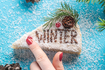 Piece of white mousse cake covered with coconut flakes imitating snow on a blue background. There is word Winter on the slice. Concept of the season winter