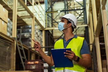 Young Indian factory warehouse worker wearing a protective face mask and safety helmet while working in logistic industry indoor. 30s man checking stock order during Coronavirus Covid 19 pandemic