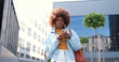 African American young stylish curly woman tapping or scrolling on smartphone and standing at city street. Beautiful female texting message on mobile phone and chatting. Outside.