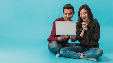 Asian young couple sitting and using laptop with smiling face together isolated on blue background.Concept of happy couple working with online technology.