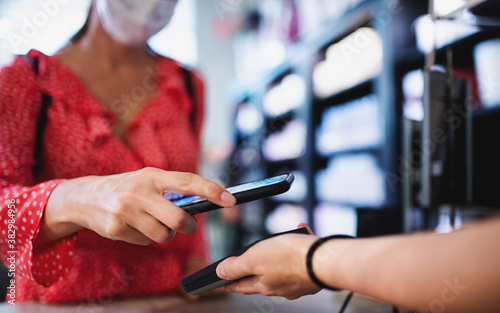 Fotografía Woman with face mask paying by smartphone indoors in shopping center, coronavirus concept