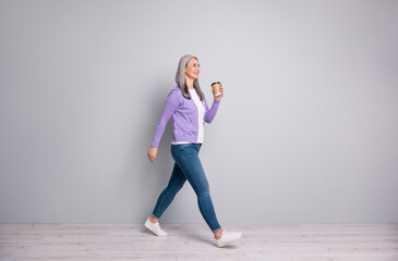 Full length body size photo of smiling elder woman having break going fast keeping cup of coffee wearing jeans cardigan eyeglasses isolated on grey color background