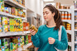 © _KUBE_ - Portrait of a young pretty Caucasian woman reads the label on a jar of olives. Shelves with product on the background. Concept of purchasing goods and shopping