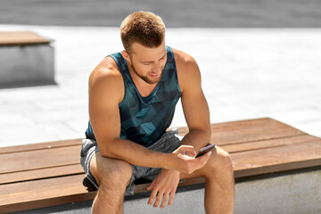 fitness, sport and technology concept - happy smiling young man with smartphone sitting on bench outdoors