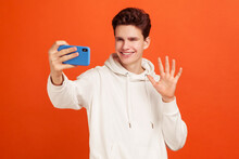 Cheerful Young Man In Stylish Hoodie Saying Hi Making Video Call Using Smartphone, Smiling With Toothy Smile, 4g Connection. Indoor Studio Shot Isolated On Orange Background