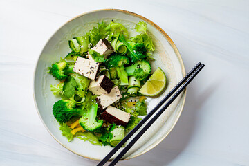 Wall Mural - Green Asian salad with broccoli and smoked tofu in white bowl, white background.