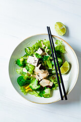 Wall Mural - Green Asian salad with broccoli, smoked tofu and peanut dressing in white bowl, white background.