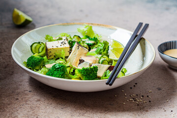 Wall Mural - Green vegan salad with broccoli, smoked tofu and tahini dressing in white bowl, dark background.