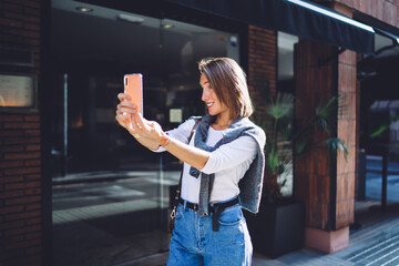 Positive caucasian female millennial holding smartphone posing for selfie on front camera on city street, smiling woman 20s making picture for share in social networks 