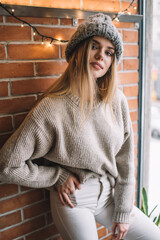 Canvas Print - Half length of fashionable Caucasian model dressed in stylish apparel looking at camera during indoors photo session, charming hipster girl in trendy clothes posing near brick wall with garland