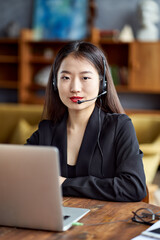 Happy asian businesswoman in headset speaking by conference call and video chat on laptop in office, smiling chinese support service agent consulting customer. Female portrait looking at camera