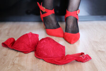 Sex Concept, Female Legs And Removed Lace Bra On A Floor. Woman In Fishnet Stockings And Red Shoes On High Heels In A Bedroom, Seductive Lingerie For Romantic Date
