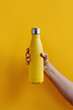 Close up of female hand, holding yellow reusable steel bottle on yellow background