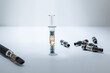 A THC distillate syringe standing upright, in the middle of used vape cartridges, and a vape pen on the other side.  Spot light, white surface and background with dramatic light.