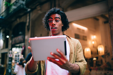 Wall Mural - Handsome curly hipster guy in eyewear reading news and stressed with updates on digital tablet, handsome young millennial male 20s watching sholing video on touchpad standing outdoors near neon lights