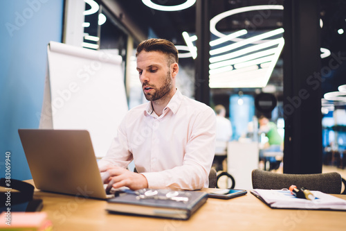 Caucasian male employee reading web information on browsed site survey during productive working process at office table, skilled businessman using 4g internet for making online banking and booking