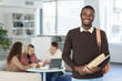 © Seventyfour - Waist up portrait of smiling African teenage boy looking at camera and smiling while standing in college library with people working in background, copy space