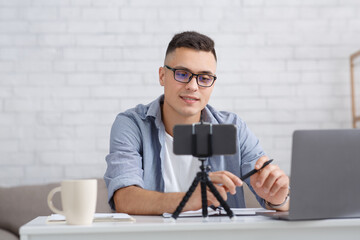 Modern blogger, teacher or tutor and technology for working at home with students. Young man with glasses chatting online in living room interior with smartphone and laptop