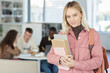 © Seventyfour - Waist up portrait of blonde young woman looking at camera and smiling while standing in college library with people working in background, copy space