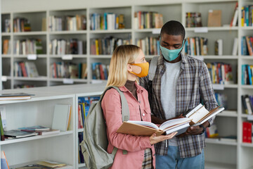 Wall Mural - Waist up portrait of two students wearing masks while standing in school library and holding books, copy space