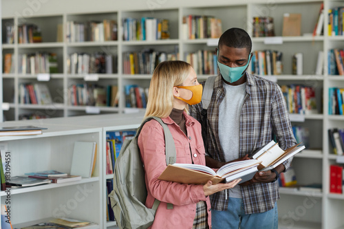 Waist up portrait of two students wearing masks while standing in school library and holding books, copy space