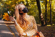 © maryviolet - Pretty young woman wears protective mask walking in autumn park holding leaves. Coronavirus covid-19 fashion