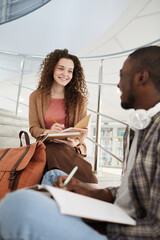 Wall Mural - Vertical portrait of two students chatting while sitting on stairs in college and working on homework, focus on curly haired young woman smiling cheerfully