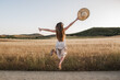 Beautiful young blonde woman with long hair on a summer afternoon jumping on her back in the field. Girl with hat in hand and white dress happy giving a big jump