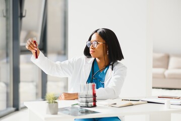 Young beautiful African American girl doctor in a white coat with a stethoscope. sitting at a table with reagent flasks on white background. Female lab assistant studying blood sample for analysis