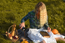 A Blond Young Woman Dressed In A Country Style Has A Picnic Sitting On Picnic Blanket With Basket, French Bread, Coffee, Pumpkins And Old Book