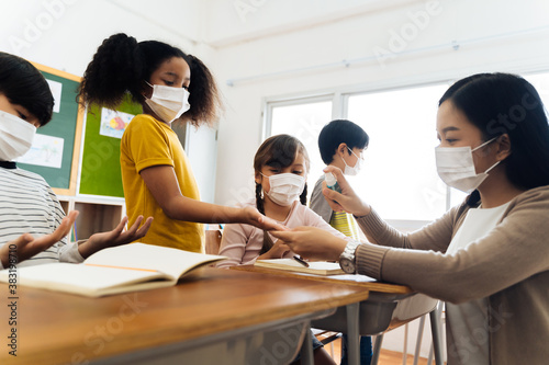 Young female teacher using an alcohol spray to disinfect student hands in classroom. Asian woman in face mask cleaning pupils\' hands with hand sanitizer. School reopen after quarantine and lockdown.