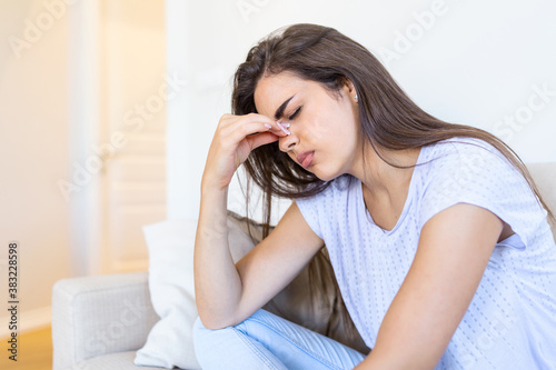 Portrait of an attractive woman sitting on a sofa at home with a headache, feeling pain and with an expression of being unwell. Upset woman sitting on couch feeling strong headache migraine.