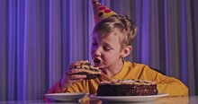 Happy Child Takes A Piece Of Celebration Cake And Then Bite It And Swallow.  Cute Boy Eats Cake On His Birthday. Happy Boy With Cone On Head Celebrates His Birthday. Caucasian Child And Birthday Cake