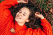 © iuricazac - Top view of a gorgeous young woman smiling and wearing orange knitted sweater posing on the grass with autumn leaves. Beautiful female with closed eyes has dreamy expression lying outdoor in the park.