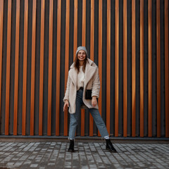 Urban happy young woman in a fashion hat in a stylish eco-coat in vintage jeans in leather black trendy boots stands and smiles near a modern building in the city. Joyful cute girl. Casual spring look