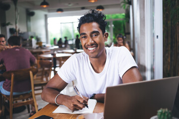 Content Hispanic student taking notes while using laptop in cafe