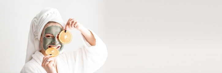 Wall Mural - cosmetology and spa. young woman having clay mud mask on her face, holding slices of grapefruit on white background. Long banner format