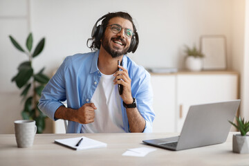 Wall Mural - Cheerful indian freelancer guy singing and listening music with headphones while working