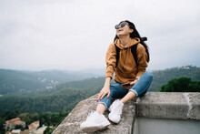 Overjoyed Asian Female Traveler In Sunglasses Enjoying Active Leisure And Destination Exploring, Smiling Woman Tourist Sitting Outdoors Satisfied With Wanderlust Tour On Discovering Locations