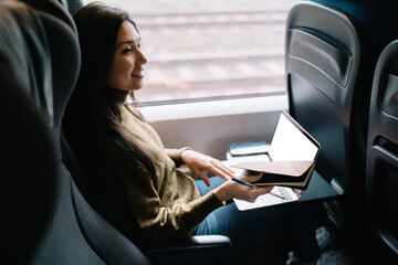 Wall Mural - Smiling creative female writer enjoying work on journey traveling by train with technology and wireless connection, prosperous business woman satisfied with online project sitting in wagon with laptop