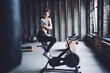 © BullRun - Young caucasian sports women in activewear sitting on exercising bike looking at smartphone choosing playlist songs, pretty female athlete looking at mobile phone on arms during workout