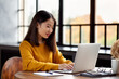 © Nata Bene - Asian woman working on laptop at home or in cafe. Young lady in bright yellow jumper is sitting at desk typing on computer. Business oriental female in front of window
