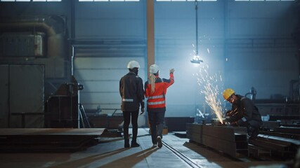 Wall Mural - Two Heavy Industry Engineers Walk Away from Camera in Steel Factory and Discuss Work. Industrial Worker Uses Angle Grinder in the Background. African American Specialist Talks to Female Technician.