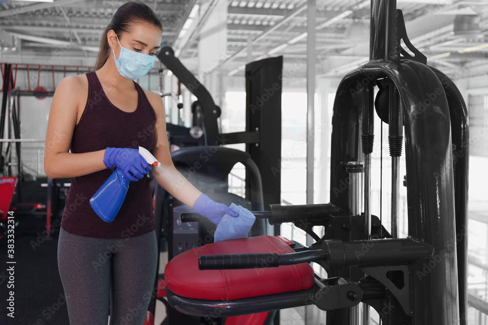 Woman cleaning exercise equipment with disinfectant spray and cloth in gym - obrazy, fototapety, plakaty 
